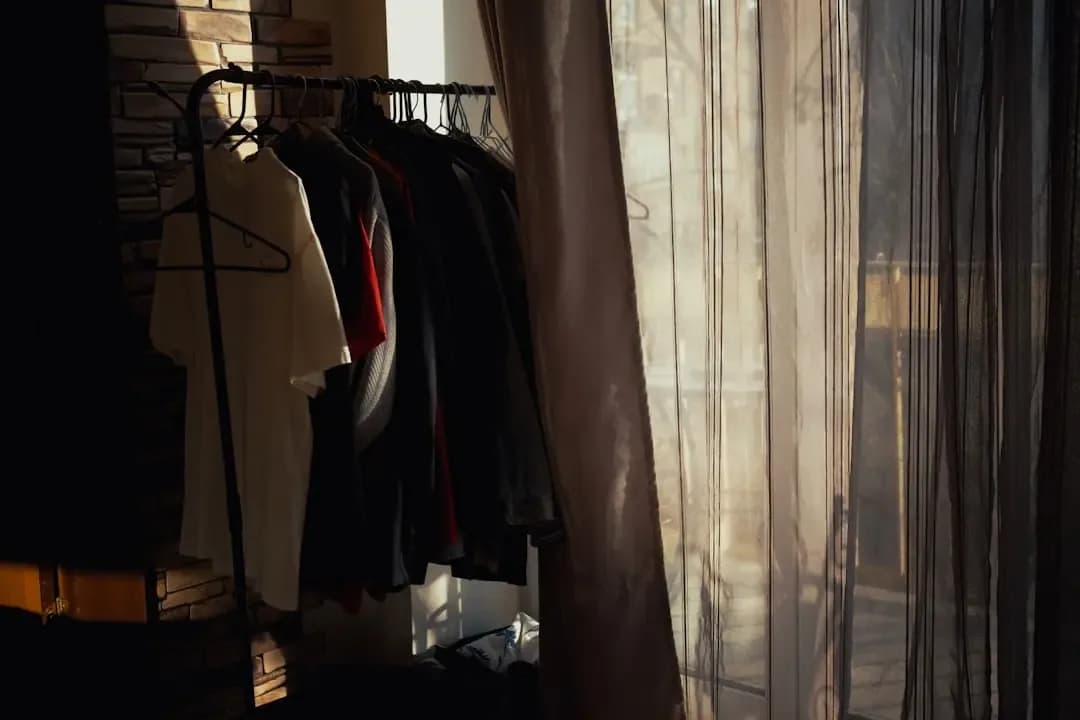 a rack of clothes next to a window with sheer curtains